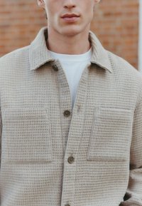 Beige textured shirt jacket with a woven pattern, closed with brown buttons, featuring two front pockets and a classic collar.