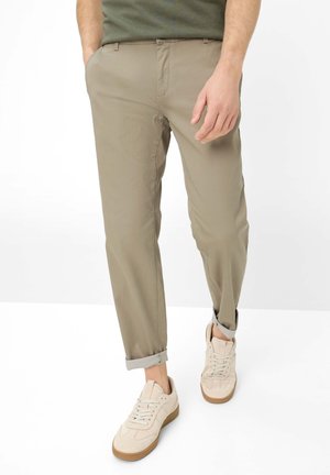 Mann trägt hochgekrempelte beige Chinos, olivgrünes Hemd und beige Sneakers, steht mit einer Hand in der Tasche vor weißem Hintergrund.