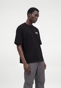 Uomo che indossa una maglietta nera oversize con testo bianco sul petto e pantaloni grigi, in piedi davanti a uno sfondo bianco uniforme.