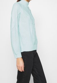 Ljust blått stickat tröja med hög krage, strukturerat mönster och ribbad manschett. Styla med svarta byxor.