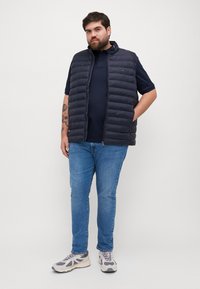 Jūras spēku pufīgs bezpiedurkņu vestes, ar rāvējslēdzēju, uzvilkta virs tumšas polo krekliņa. Piegriezums ir pieguļošs, viegls audums; apvienots ar ziliem džinsiem un pelēkiem sporta apaviem.