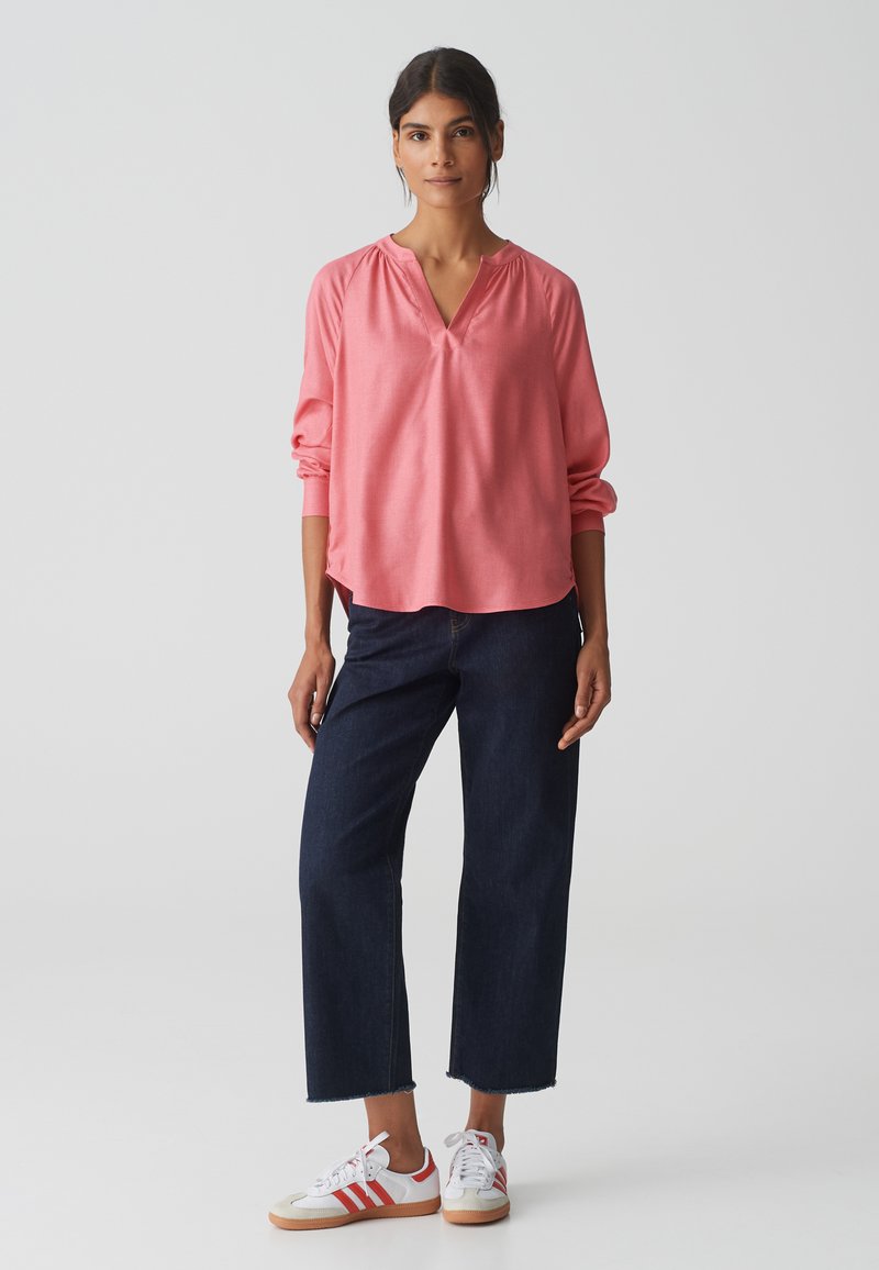 Top rosa chiaro a maniche lunghe con scollatura a V e dettagli arricciati, abbinato a jeans cropped blu scuro e sneakers bianche con strisce rosse.