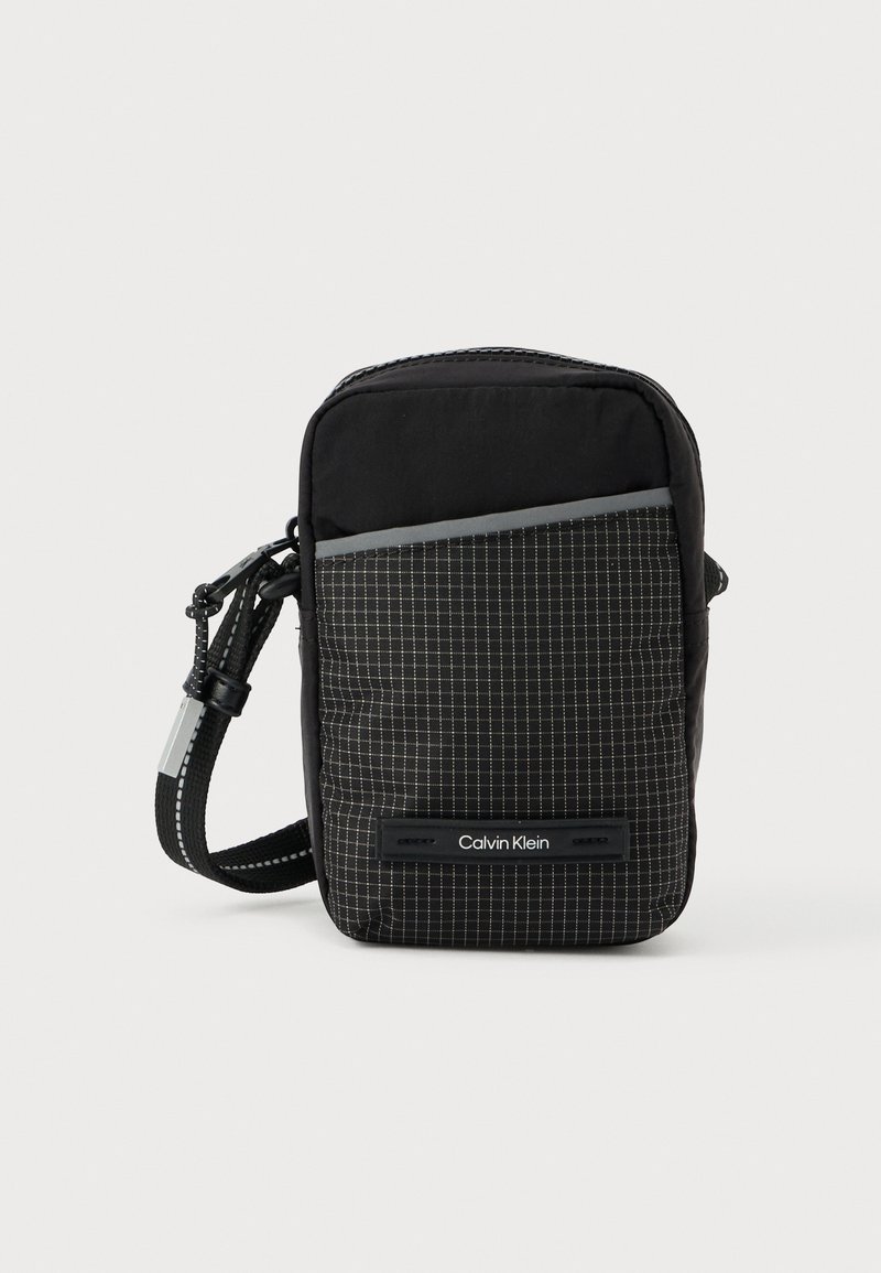 Fekete, kockás mintájú keresztpántos táska, texturált anyagból készült. Elülső zsebbel és logó díszítéssel. Állítható pánttal ellátva.