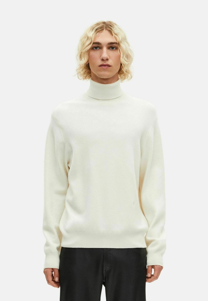 Suéter de cuello alto crema con textura suave, de corte holgado y puños acanalados. Combinado con pantalones negros, contra un fondo blanco liso.