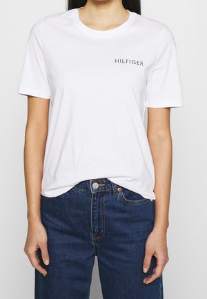 Woman wearing a plain white short-sleeve t-shirt with "HILFIGER" text near the left shoulder, paired with blue denim jeans.
