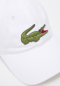 Biała czapka z zakrzywionym daszkiem, z zielonym logo krokodyla wyszytym z przodu, z czerwonymi detalami w okolicy jego pyska.