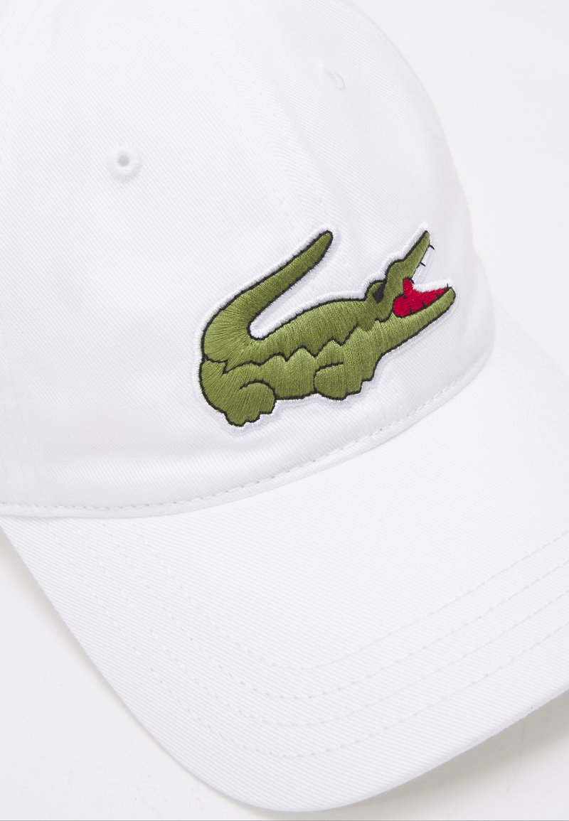 Gorra blanca con visera curva que presenta un logo de cocodrilo verde bordado en la parte frontal, con detalles rojos en su boca.