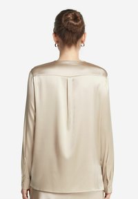 Satinbeige blus med rund nackdesign bak, långa ärmar, knäppta manschetter och en mitt bak-veck för extra designdetalj.