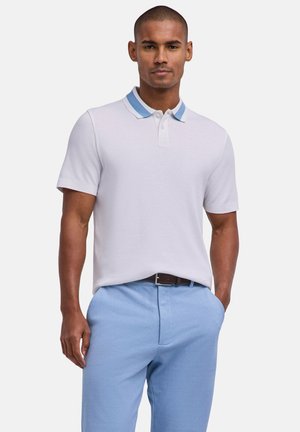 Uomo che indossa una polo bianca a maniche corte con colletto blu e pantaloni azzurri, in piedi con una mano in tasca.
