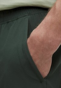 Dunkelgrüne Sportshorts mit glatter Oberfläche, elastischem Bund und Seitentaschen. Fokus auf den Stoff und funktionale Designelemente.