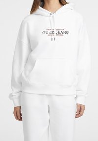 Sudadera blanca con capucha hecha de mezcla de algodón, que incluye un bolsillo frontal, capucha con cordón y logo impreso en negro y rojo.