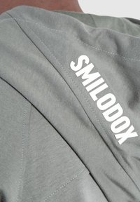Šedá mikina s hladkou strukturou, diagonálními švy a textem "SMILODOX" v tučných bílých písmenech na zádech.