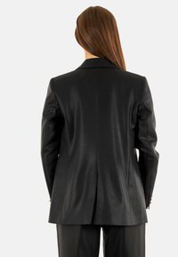 Blazer nero su misura con una texture liscia, spacco singolo sul retro e bottoni lucidi sulle maniche. Visto da dietro.