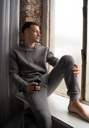 Jonge man in een grijze gewatteerde hoodie en broek, die een zwarte mok vasthoudt terwijl hij blootsvoets op een vensterbank zit en naar buiten kijkt door een gestructureerd raam.