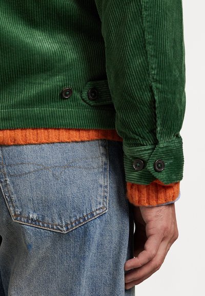 Giacca in corduroy verde con polsini arancioni a coste, dotata di bottoni neri e una superficie texturizzata, indossata sopra jeans di denim blu con una tasca visibile.