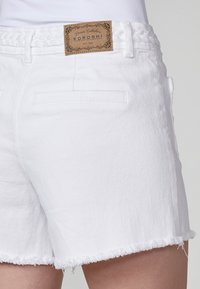 Bermudas de mezclilla blanca con dobladillo deshilachado, detalle de cinturilla trenzada y bolsillos traseros. Etiqueta de la marca "KOROSHI" unida a la cinturilla.