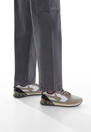 Person, die graue weiter Hosen und beigem, weißem und schwarzem Sneakers mit einer Camouflage-musternden Sohle trägt und auf einer weißen Fläche steht.
