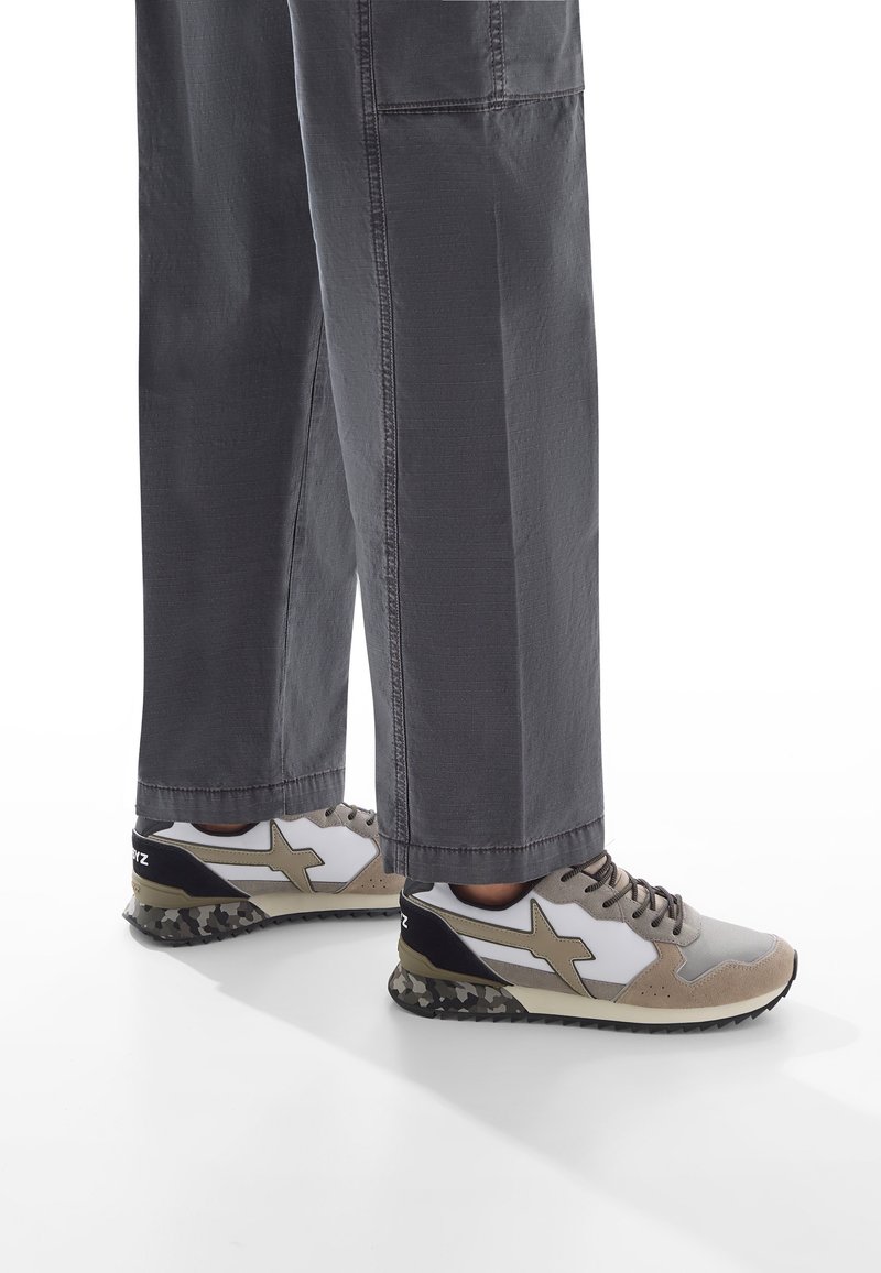 Persona che indossa pantaloni larghi grigi e sneakers beige, bianche e nere con una soletta a motivo mimetico, in piedi su una superficie bianca.