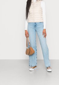 Gilet imbottito beige sopra una maglietta bianca a maniche lunghe con coste, abbinato a jeans larghi azzurri e sneakers marroni. Presente una piccola borsa a tracolla marrone.