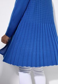 Largo cárdigan de punto azul con diseño de ribetes texturizados, mangas largas y un dobladillo acampanado, combinado con pantalones blancos.