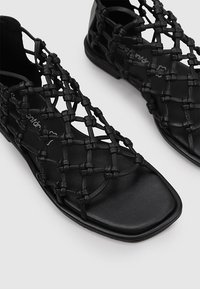 Sandalia negra con parte superior de cuero trenzado, punta cuadrada y suela plana. Presenta un intrincado patrón de nudos que proporciona ventilación y estilo.