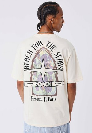 Camiseta de algodón crema con un gran gráfico que presenta patrones florales en colores pastel, con texto negro que dice "Alcanza las Estrellas" y "Project X Paris".