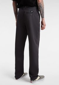 Pantalons noirs en tissu lisse, dotés d'une poche arrière avec un bouton et d'une petite étiquette de marque. Portés avec des chaussures à enfiler à carreaux.