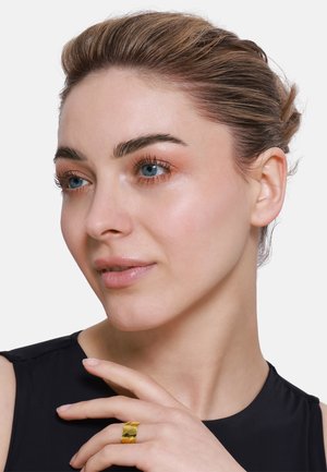 Goldfarbener, breiter Ring, der am Finger getragen wird; glatte Textur, geometrisches Design. Das Modell hat einen natürlichen Makeup-Look zu einem schwarzen Oberteil.