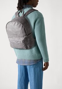 Gray backpack with smooth texture, rounded shape, front pocket, and adjustable straps. Worn over a teal knitted sweater and layered shirt.
