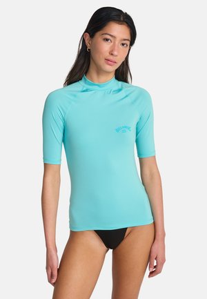 Kobieta ubrana w turkusową koszulkę z krótkim rękawem i czarne bikini, stojąca na tle jednolitego białego tła.