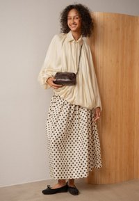 Donna sorridente, che indossa una blusa beige ampia, una gonna lunga a pois, ballerine nere, e tiene in mano una piccola borsa a tracolla marrone scuro, mentre è in piedi dentro a un ambiente.