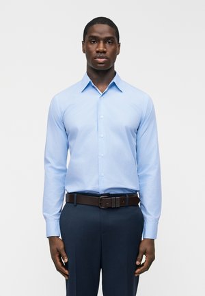 Moški, oblečen v svetlo modro srajco z gumbi, zatlačeno v temne hlače s temno rjavim pasom, stoji pred enostavnim ozadjem.