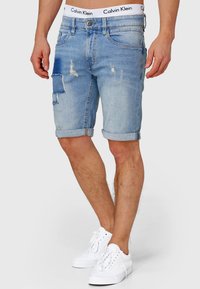 Shorts en denim bleu avec une finition délavée, des détails usés et un patch sur la jambe gauche. Les ourlets sont retroussés et le logo sur la ceinture blanche est visible.