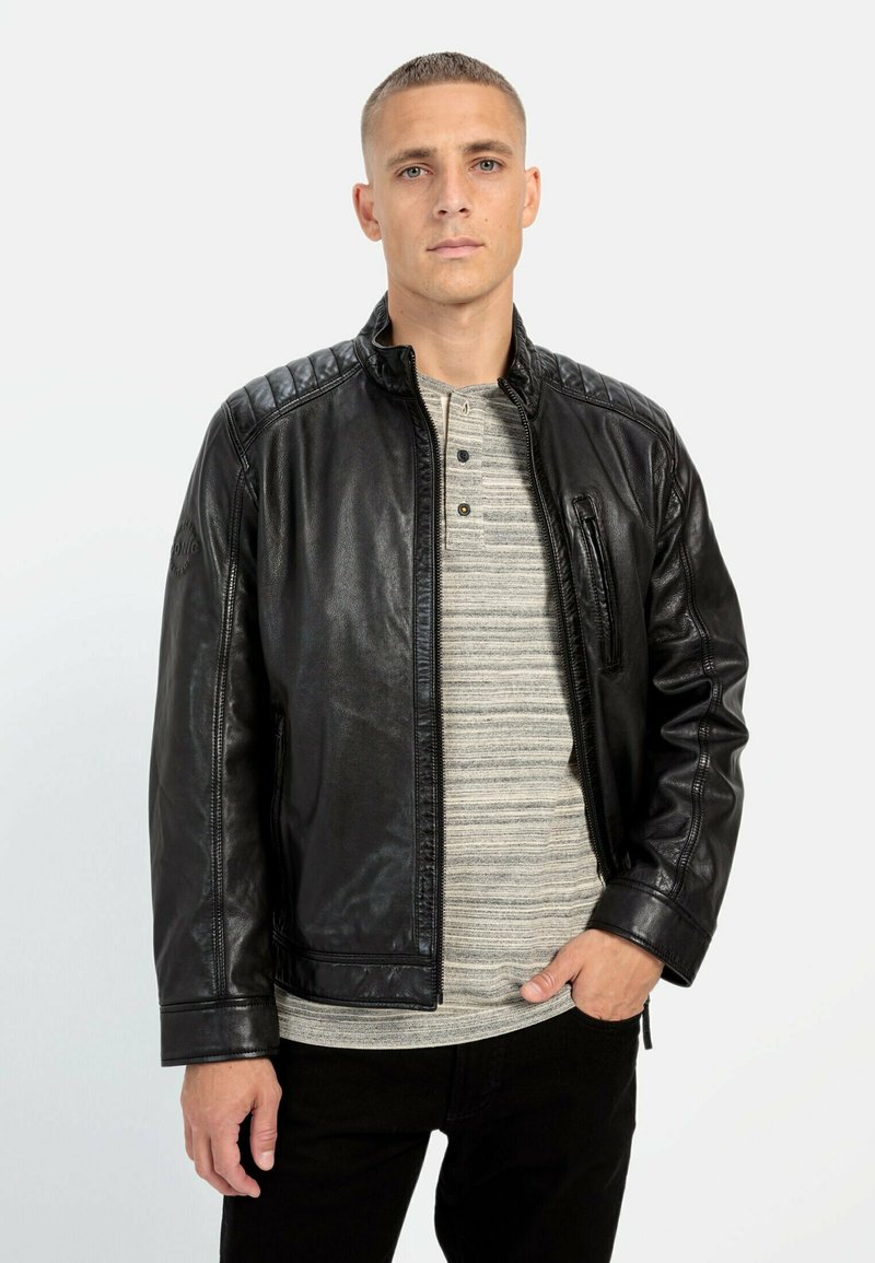 Sort læderjakke med quilted skulderdetalje, frontlynlås og en brystlomme, båret over en grå stribet henley-skjorte og sorte bukser.