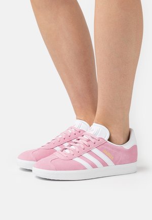 Sneakers in suede rosa con lacci bianchi e tre righe bianche. Presentano una suola in gomma e un marchio laterale in giallo. Design classico.