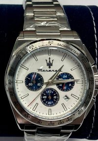 Silverfärgad armbandsur i rostfritt stål med vit urtavla, blå subskivor och tachymeter bezel. Utrustad med länkarmband och en logotyp av varumärket längst upp.