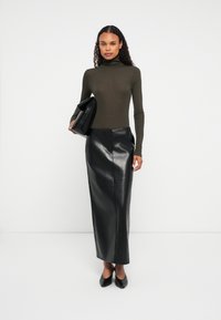 Kobieta w ciemnozielonym golfie, czarnej skórzanej spódnicy midi i czarnych szpilkach trzymająca dużą czarną kopertówkę na białym tle.