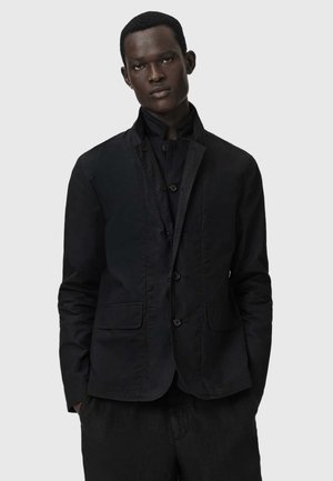 Mężczyzna o ciemnej karnacji, ubrany w czarną zapinaną marynarkę i czarną warstwową koszulę, stojący na tle gładkiego jasnoszarego tła.