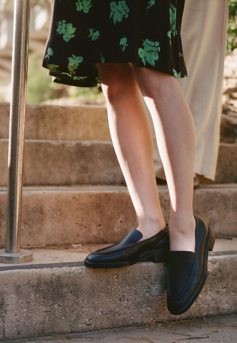 Czarne skórzane buty wsuwane o gładkim wykończeniu, z lekko podniesionym obcasem i okrągłym czubkiem, umieszczone na kamiennych schodach obok kwiecistej sukienki.