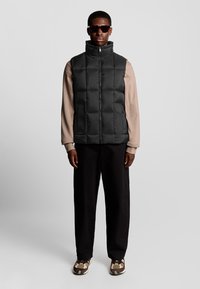 Schwarze gepolsterte Weste mit hohem Kragen, Reißverschluss und Seitentaschen, getragen über einem beigen Langarmshirt und schwarzen Hosen. Kombiniert mit Sonnenbrille und gemusterten Schuhen.