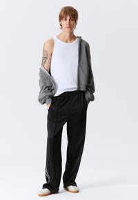 Grau zip-up Kapuzenpulli über einem weißen Tanktop, kombiniert mit schwarzen Sporthosen mit weißen Seitenstreifen und beigen Sneakers.