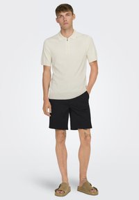 Cremefarvet polo med korte ærmer og lynlås-krave, parret med sorte shorts og brune slip-on sandaler. Modellen har tatoveringer på begge ben.
