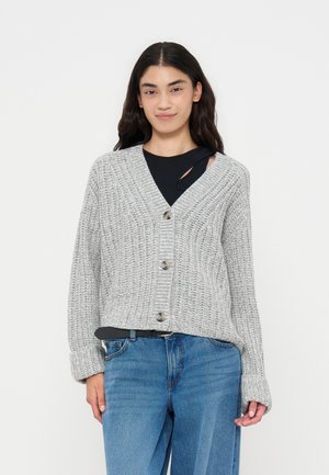Kvinde iført en lysegrå knappet strikket cardigan over en sort top og blå jeans, stående mod en ensfarvet hvid baggrund.