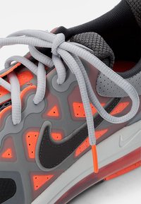Chaussure de sport grise et orange avec le logo Nike noir, lacets gris noués en nœud et doublure intérieure noire texturée.