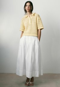 Mujer que lleva un polo amarillo de punto de manga corta, pantalones blancos de pierna ancha y cintura alta, y sandalias blancas abiertas, de pie contra una pared lisa.