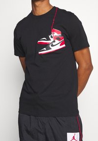 Camiseta negra con un gráfico de zapatillas rojas y blancas en el pecho. Manga corta, cuello redondo, fabricada en algodón.