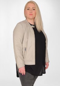 Ljusbeige imitationsläderjacka med rund krage, dragkedjefickor på sidorna och en figursydd design, bärs över en svart topp, kombinerad med mörka jeans.