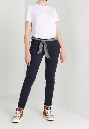 Biała koszulka z krótkim rękawem w połączeniu z granatowymi spodniami, ozdobiona paskiem w cętki. Model nosi pomarańczowo-białe trampki z wysoką cholewką.