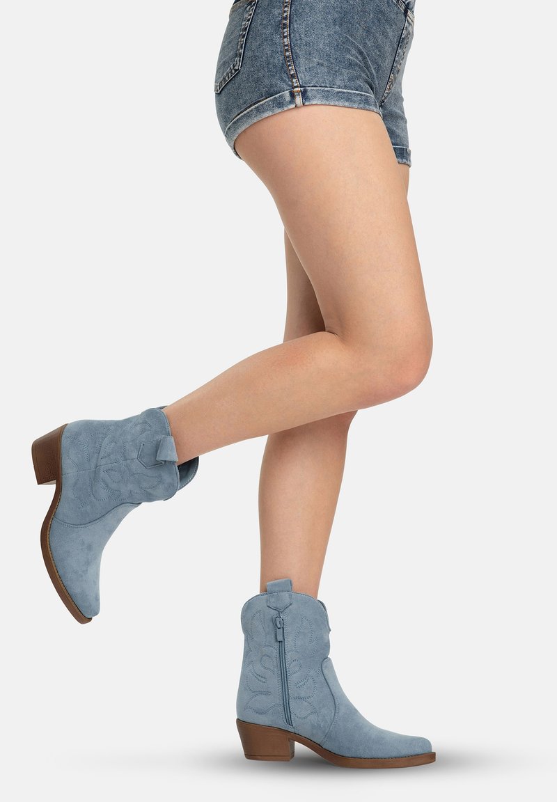 Benenden in denim shorts en lichtblauwe suède mid-calf laarzen met lage hakken en zijritsen, één been naar achteren gebogen.
