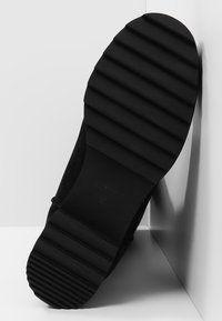 Černá semišová bota s vroubkovou gumovou podrážkou. Vyznačuje se hladkým, zakřiveným tvarem a minimálními detaily. Boční pohled zvýrazňuje texturu podrážky.